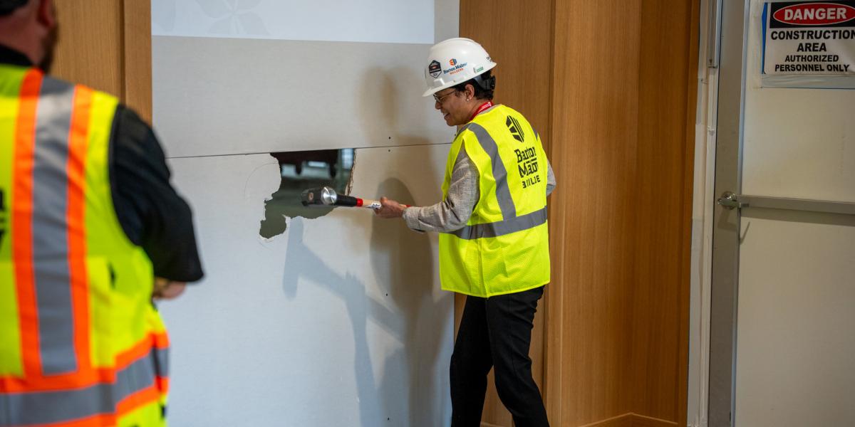 A woman is seen with a large hammer while wearing a hard hat and neon yellow work vest.