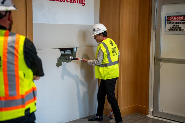 A woman is seen with a large hammer while wearing a hard hat and neon yellow work vest.